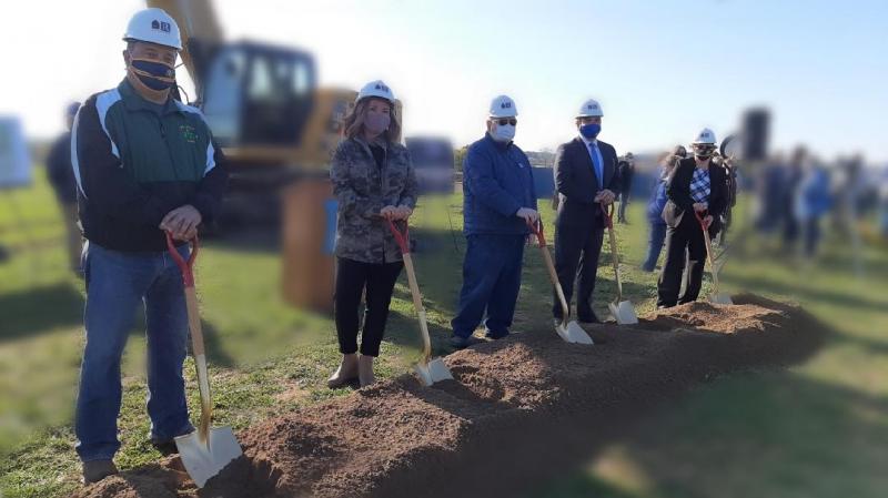 The five Board of Education members are lined up horizontally in front of a two-foot wide, one-foot deep, 15-foot long mound of soil. They have white hard hats on and shovels stuck in the dirt. A podium, display easels and a piece of construction machinery are in the background.They are spaced roughly three feet apart and wearing masks in observation of COVID-19 protocols.