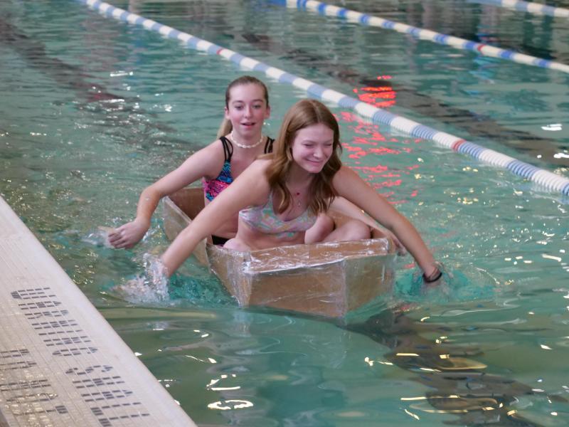 Students sit in and paddle a self-made cardboard boat in a lane of a pool.