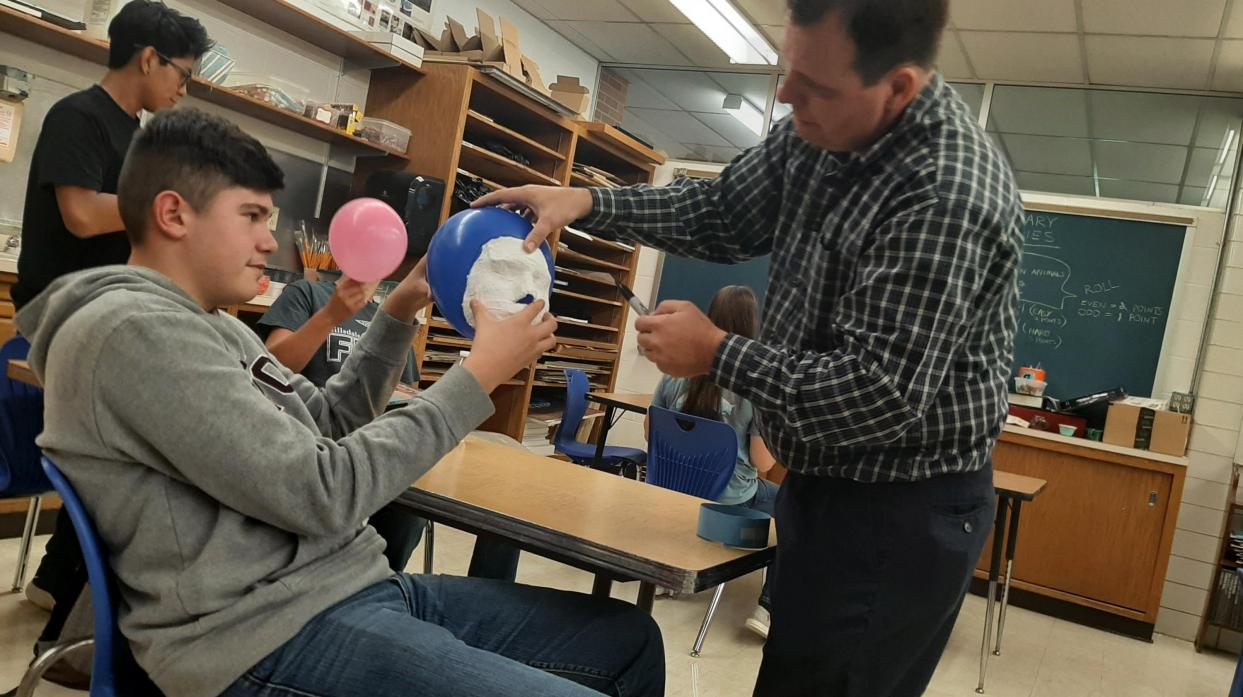 A student holds an inflated balloon and face mask while a teacher outlines the mask on the balloon.