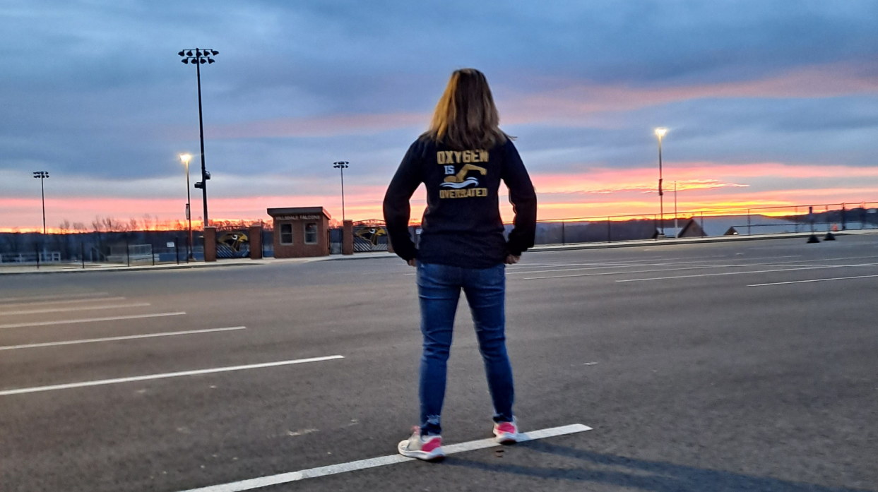 A swimmer poses in the school parking lot facing the sunrise. The back of her jacket reads "Oxygen is overrated."