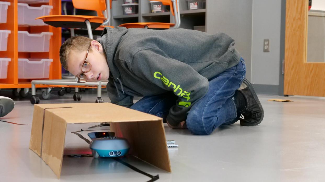 A student looks through a cardboard tunnel as a finchbot follows a black line through it.