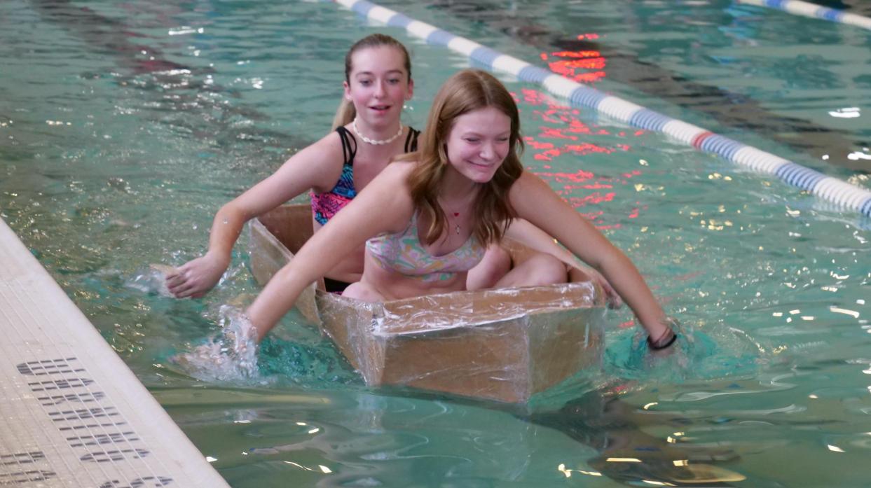 Students sit in and paddle a self-made cardboard boat in a lane of a pool.