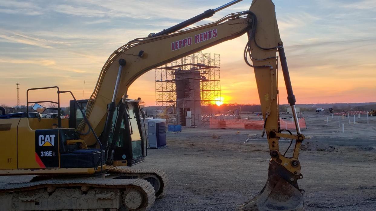 The raised arm of an excavator frames the elevator shaft encased in scaffolding with the rising sun in shades of orange and yellow in the background.