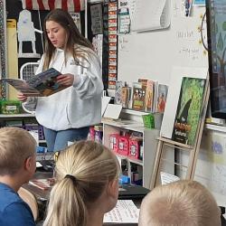 A high school student-athlete reads out loud to elementary students in their classroom.