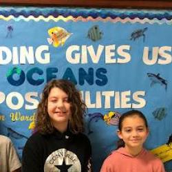 Four students stand in front of a bulletin board with the text: "Reading gives us oceans of possibilities."