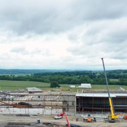 This is a drone shot looking straight on at the front of the educational building under construction in the background with part of the existing facility parking lot in the foreground.