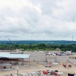 An overhead drone shot from the back of the existing rear parking lot, where the roofs of two buses are visible in the lower left corner. in the background, the elementary wing under construction is visible in the top left corner with foundation work for other parts of the building filling the rest of the picture to the right.