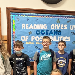 Ten students stand in front of a bulletin board with the text: "Reading gives us oceans of possibilities."
