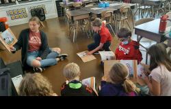 A high school student-athlete reads out loud to a small group of elementary students in their classroom.