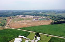 This is a bird's eye overhead drone photo from the rear of the site with a neighboring farm in the foreground, the school construction site in the middle of the photo and others' properties in the background. 
