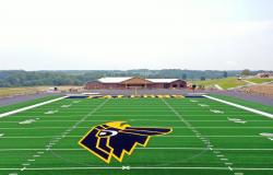 This is a drone photo looking straight on to the front of the field house in the background from the 35-yard line on the football field with the falcon head logo in the foreground.