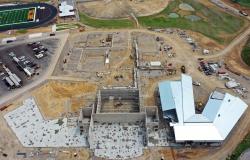 This is an overhead drone photo of the new building from the front with the poured cement of the first floor of the 7th-12th wing to the left and offices in the center, the steel roof decking of the elementary wing to the right, and the concrete block walls in the center of the foreground. In the background, the foundation work for the rest of the building can be seen with the baseball/softball complex partially pictured behind it, part of the football field to left, and secondary unpaved driveway to right.